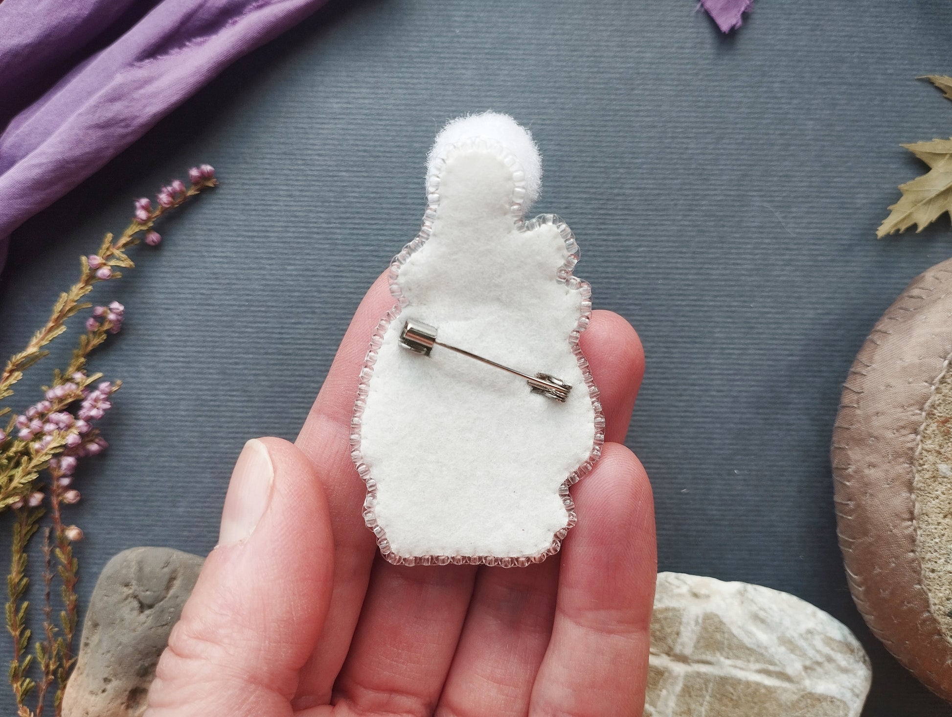 A hand holding a white felt rabbit pin against a dark background with various objects, including flowers and rocks.