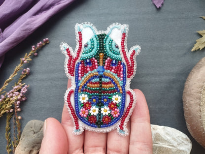 A hand holding a colorful, intricately beaded embroidered object resembling a butterfly or dragon-like creature.