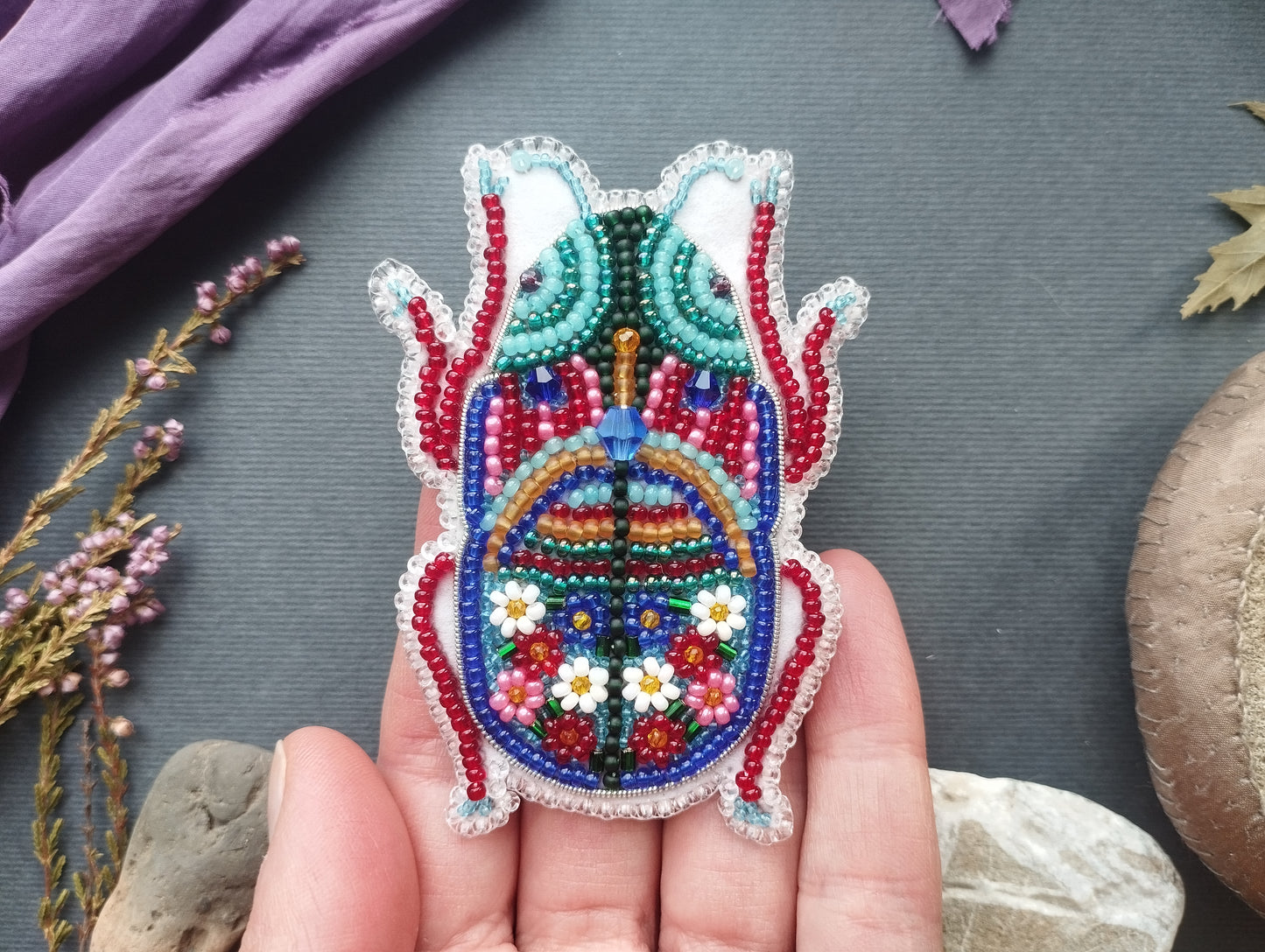 A hand holding a colorful, intricately beaded embroidered object resembling a butterfly or dragon-like creature.