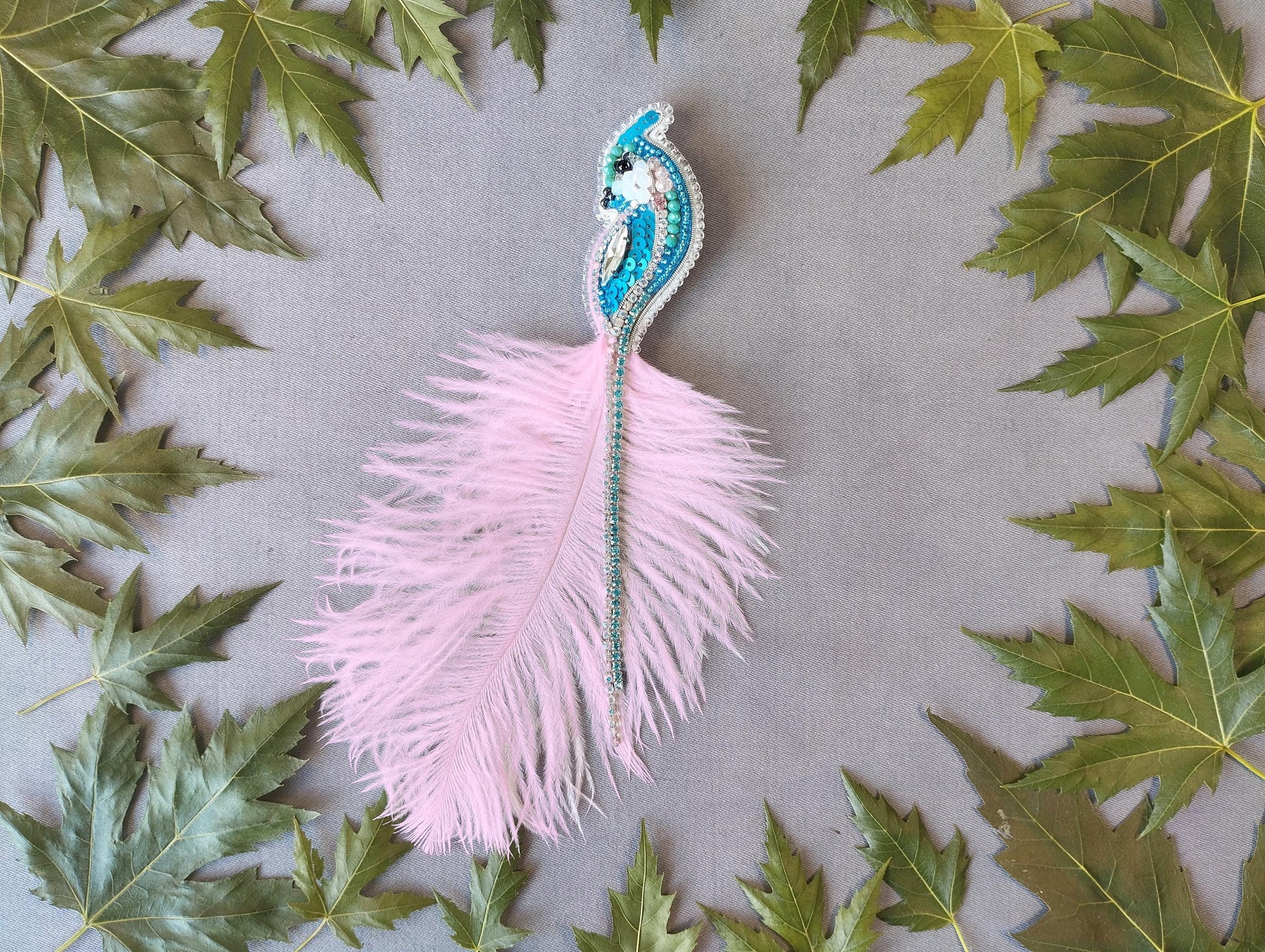 a pink feather with a blue and green beaded design, surrounded by green leaves on a gray background.