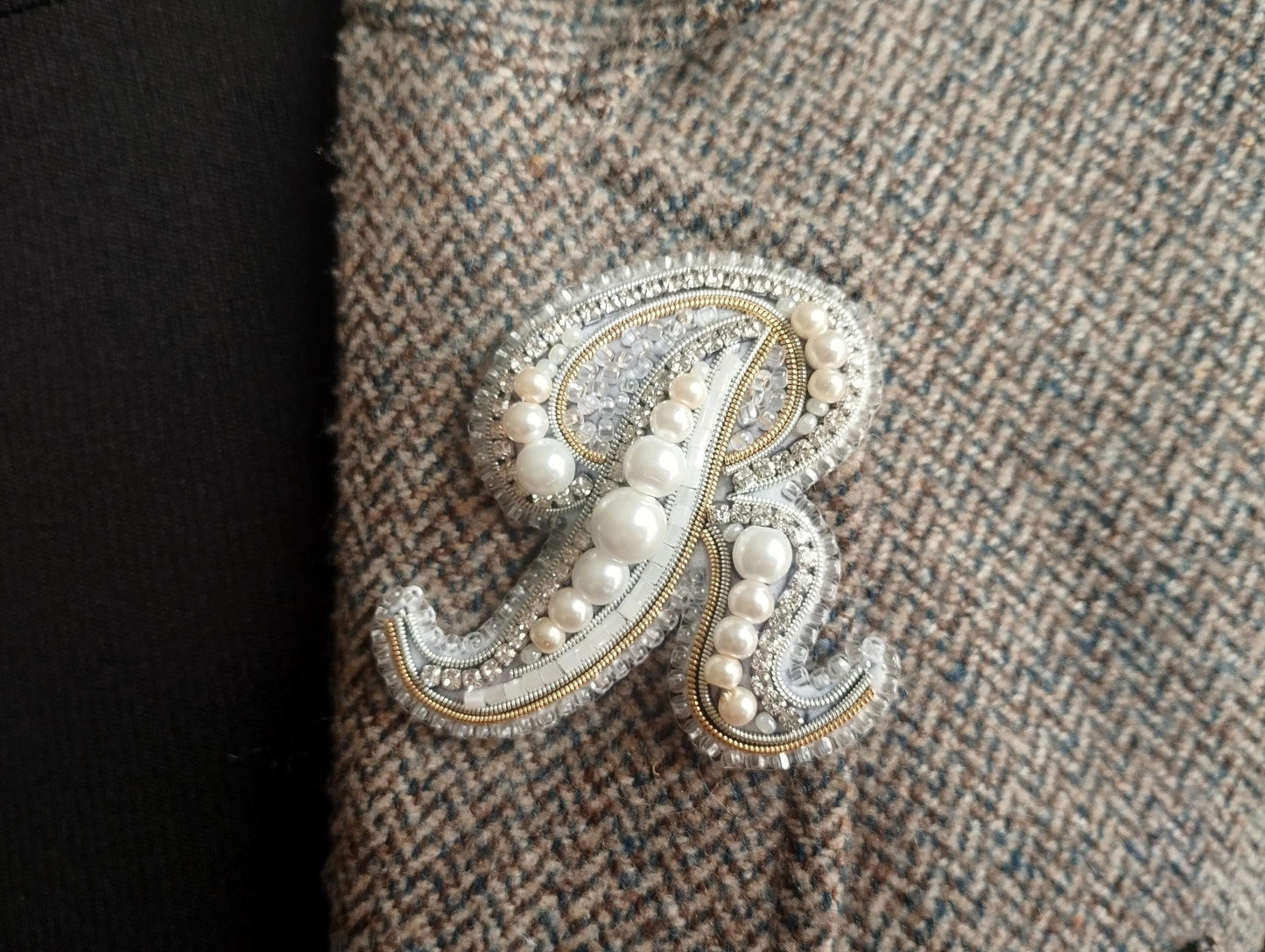 A close-up image of a decorative monogram made of pearls and beads, with a dark fabric background.