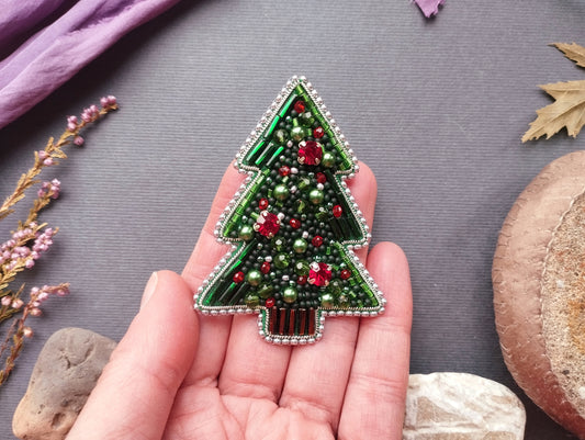 A hand holding a decorative Christmas tree brooch made of green beads and red and green beads, with a purple fabric and some leaves in the background.
