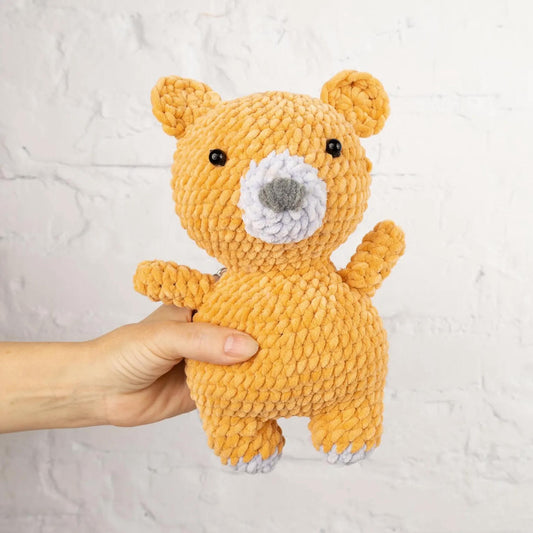 A hand is holding a small, crocheted teddy bear in front of a white brick wall.