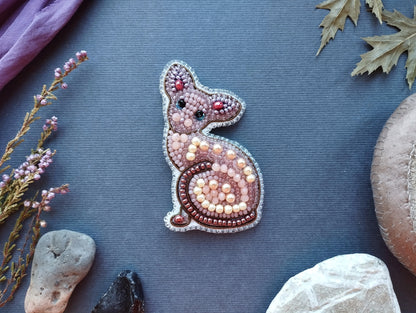 a decorative piece of jewelry, specifically a cat-shaped brooch, made of beads and pearls. The brooch is placed on a blue background, surrounded by various natural elements such as leaves, rocks, and flowers.