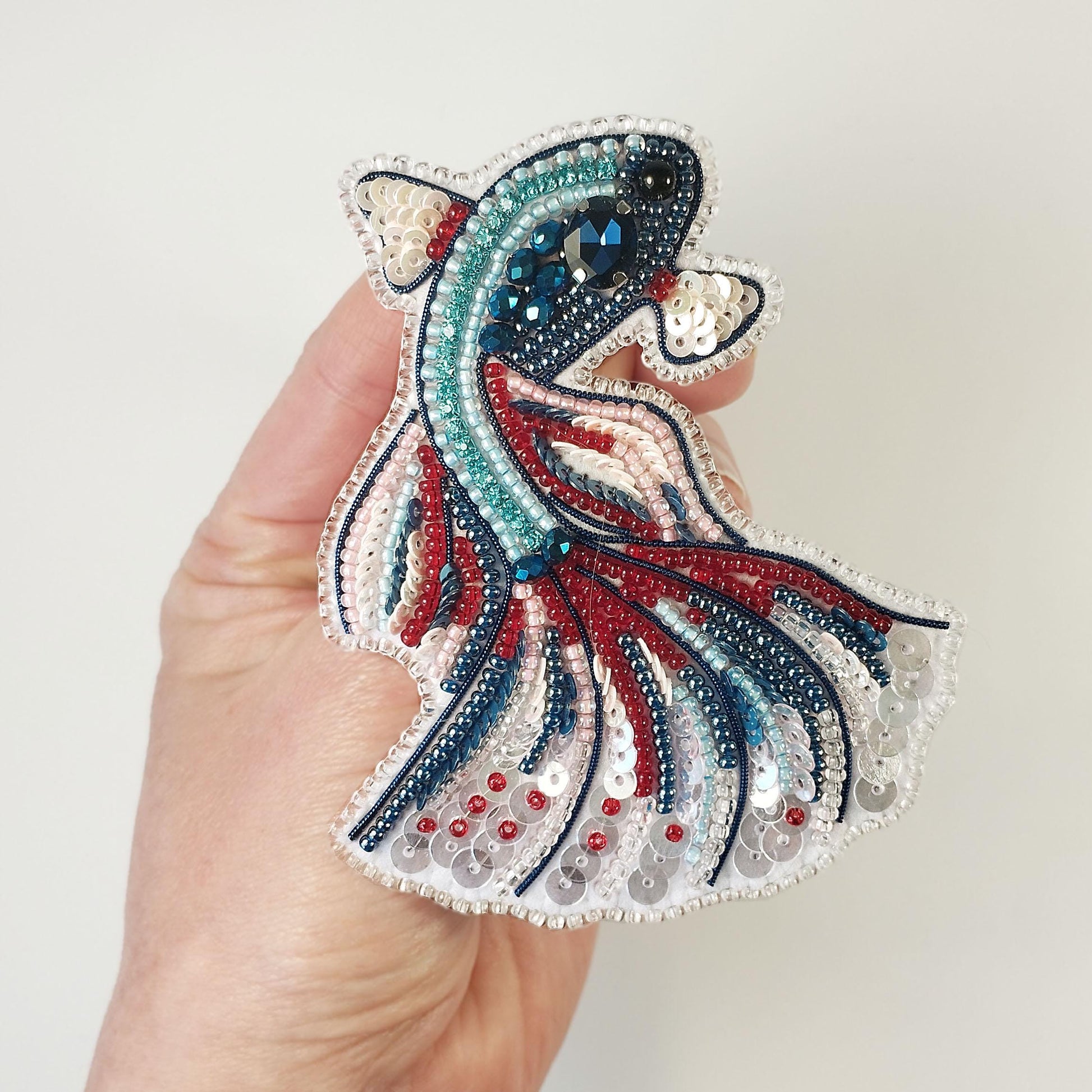 A hand holding a decorative piece of embroidered or beaded fabric featuring a fish-like design with red, blue, and white colors.