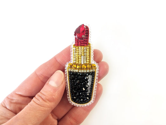 A hand holding a decorative pin or brooch featuring a red lipstick design with rhinestones and a gold base.