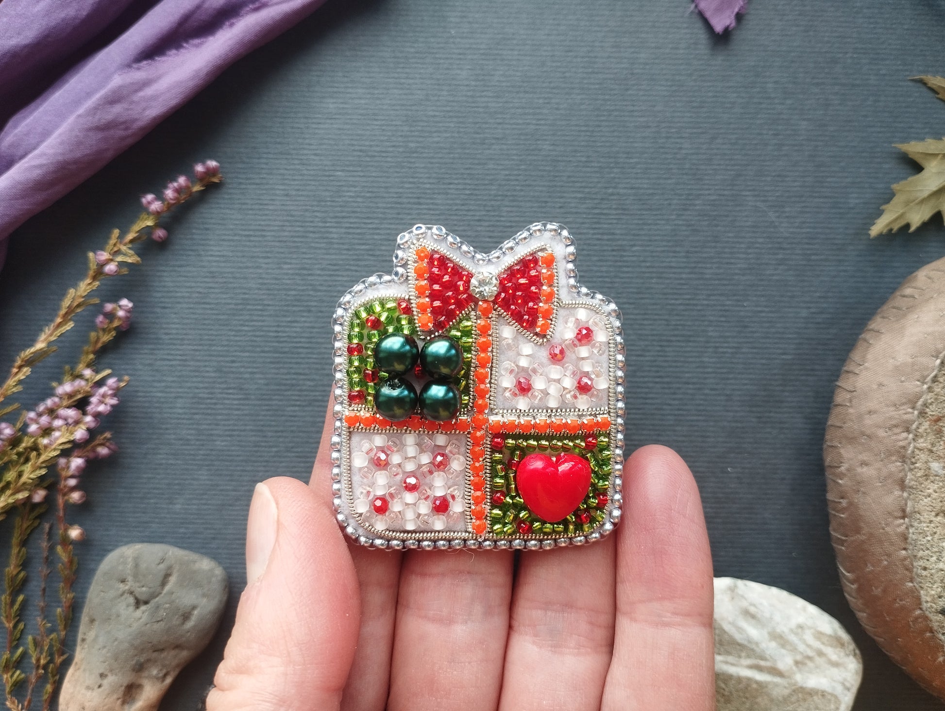 A hand holding a decorative beaded brooch with a heart-shaped button and a bow, surrounded by various natural elements such as flowers and rocks.