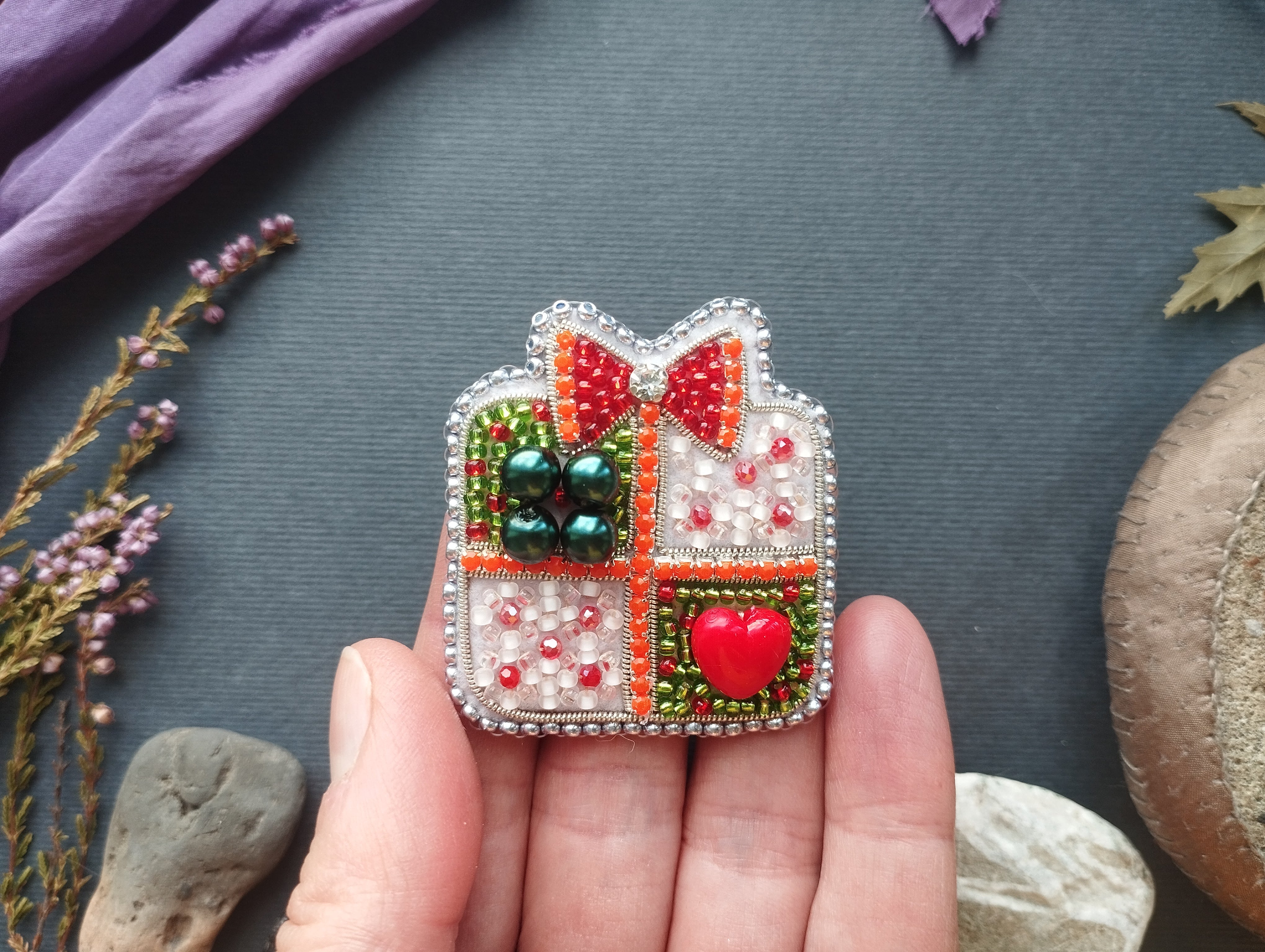A hand holding a decorative beaded brooch with a heart-shaped button and a bow, surrounded by various natural elements such as flowers and rocks.