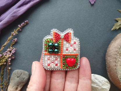 A hand holding a decorative beaded brooch with a heart-shaped button and a bow, surrounded by various natural elements such as flowers and rocks.