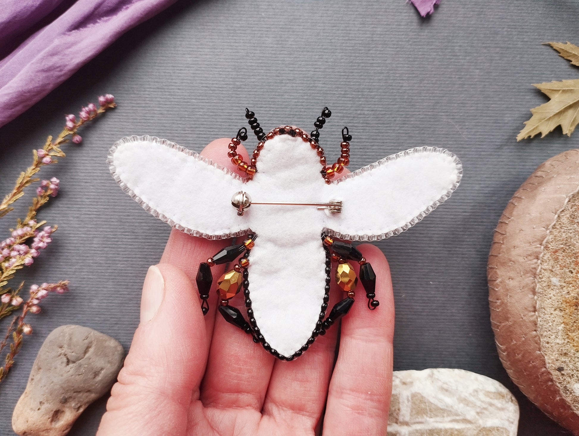 A hand holding a white felt insect pin adorned with black and gold beads, surrounded by various natural elements such as leaves, flowers, and rocks.