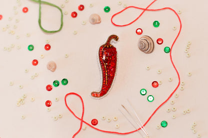 a close-up view of a red pepper embroidered with jewels, surrounded by various craft supplies such as beads, string, and buttons.