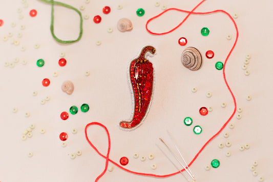 a close-up view of a red pepper embroidered with jewels, surrounded by various craft supplies such as beads, string, and buttons.