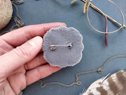 A hand holding a small gray pin cushion with a silver pin attached to it, surrounded by various accessories such as glasses, a chain, and feathers.
