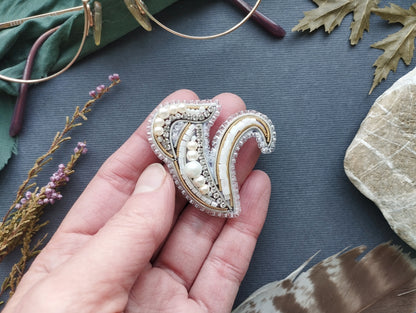 A hand holding a decorative brooch with pearls and a floral design, surrounded by various natural elements such as leaves, feathers, and a pair of glasses.