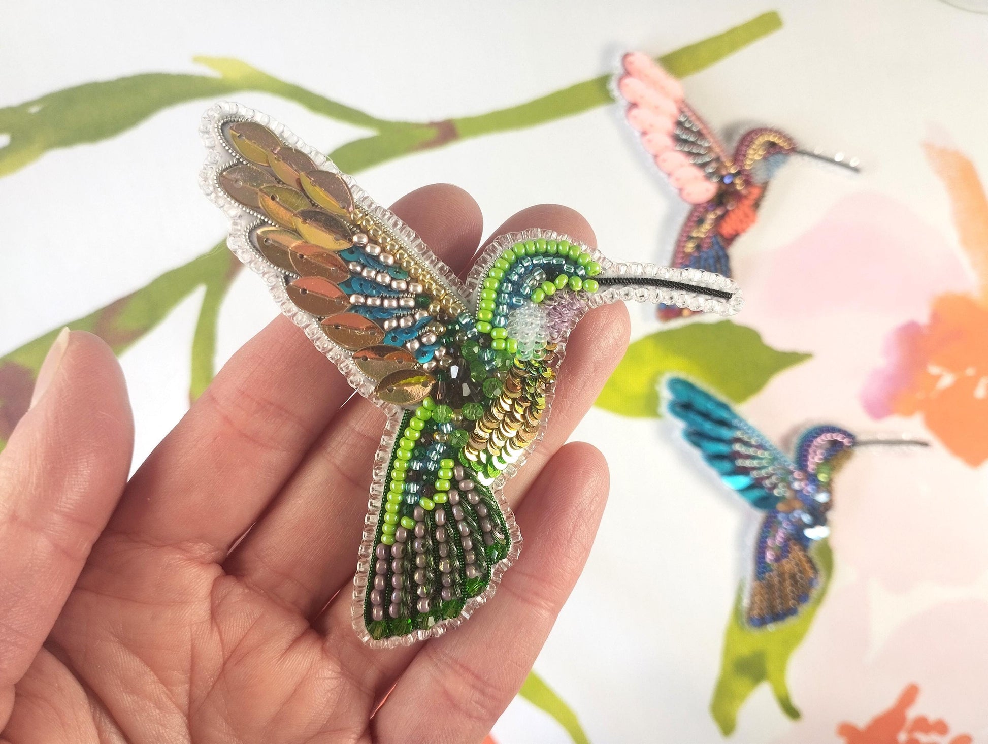 a hand holding a small brooch with a hummingbird on it
