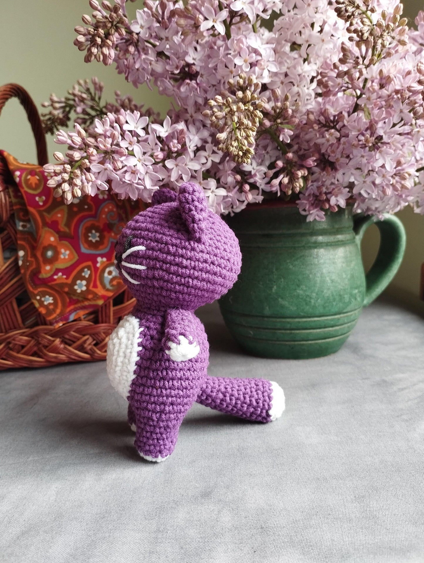 a crocheted purple teddy bear sitting next to a vase of lilacs