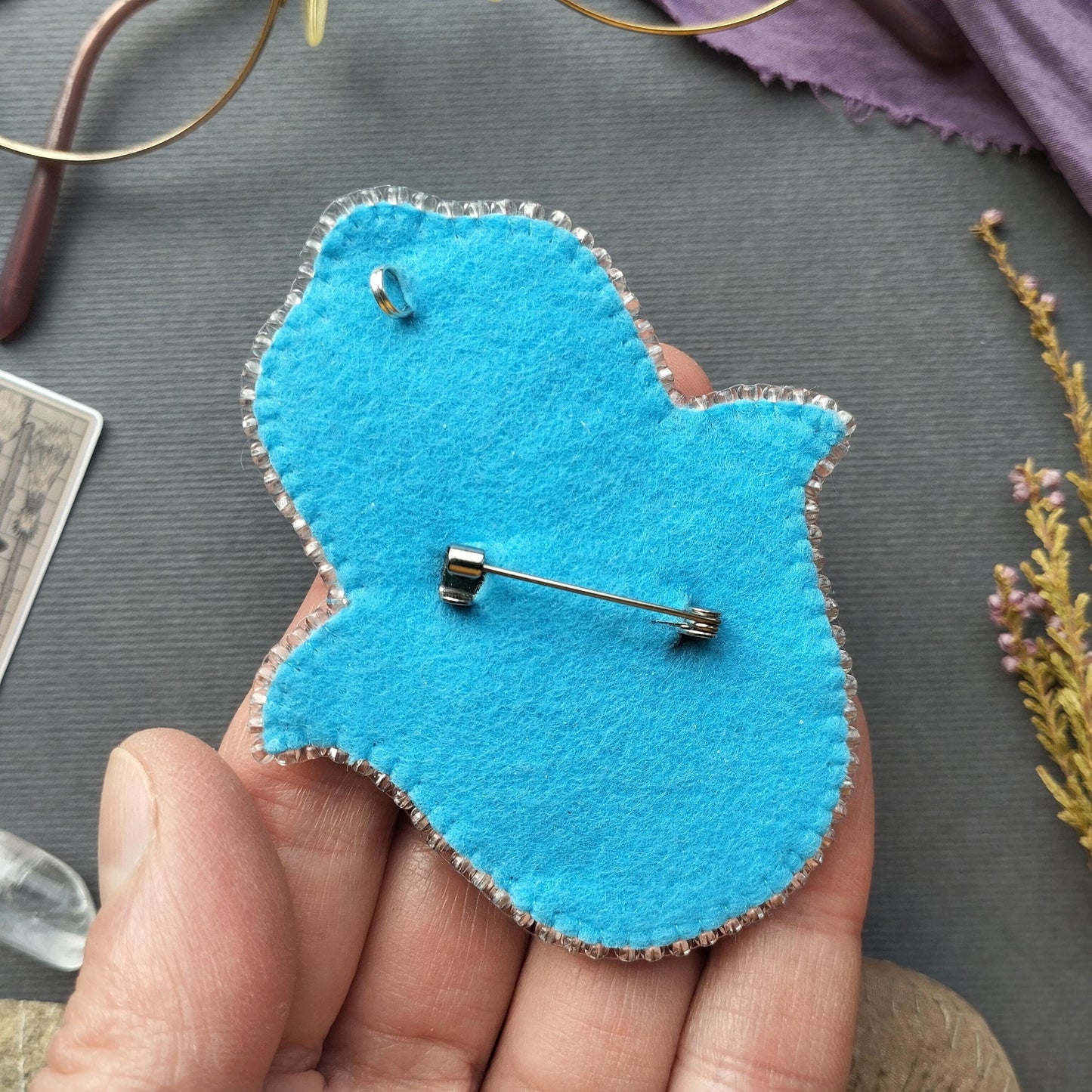A hand holding a blue felt object with a pin attached to it, placed on a gray surface.