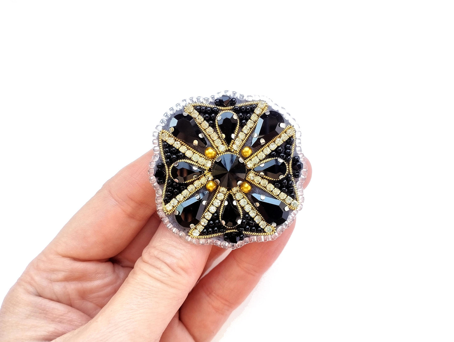 A hand holding a decorative piece of jewelry, which appears to be a ring or brooch. The jewelry is adorned with a floral design and features a combination of black and gold elements, including gemstones.