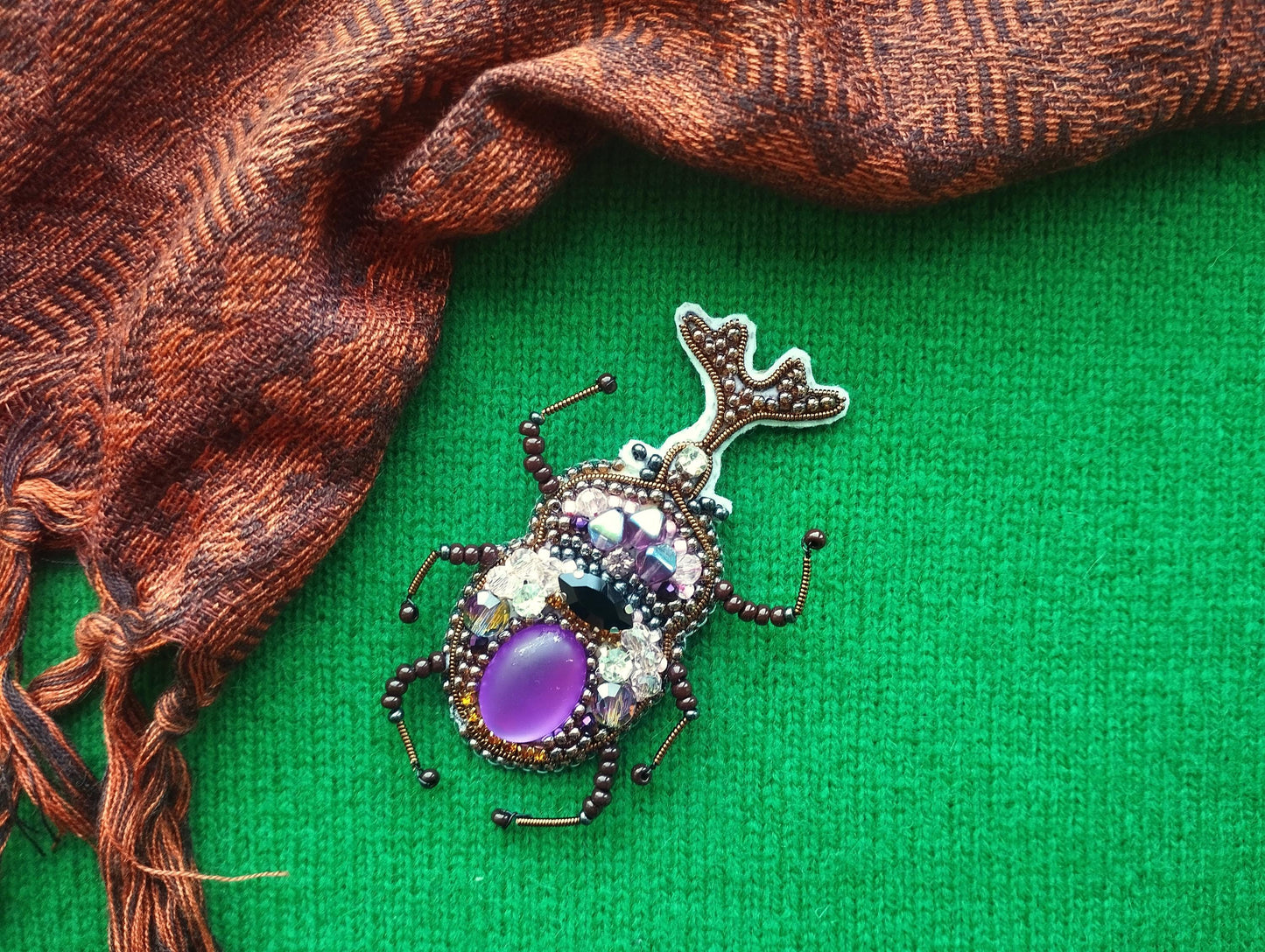 A decorative beaded insect brooch with a purple gemstone is displayed on a green background, accompanied by a brown and orange patterned fabric.