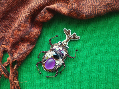 A decorative beaded insect brooch with a purple gemstone is displayed on a green background, accompanied by a brown and orange patterned fabric.