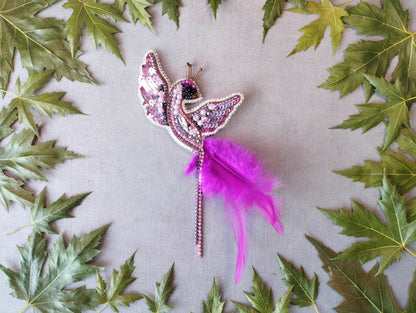 a decorative butterfly brooch with a long feather attached to it, surrounded by green leaves.