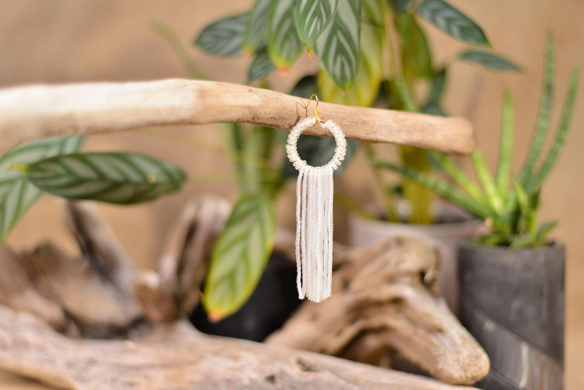 A pair of white tassel earrings with a circular design hangs from a wooden branch, surrounded by various plants and natural elements.