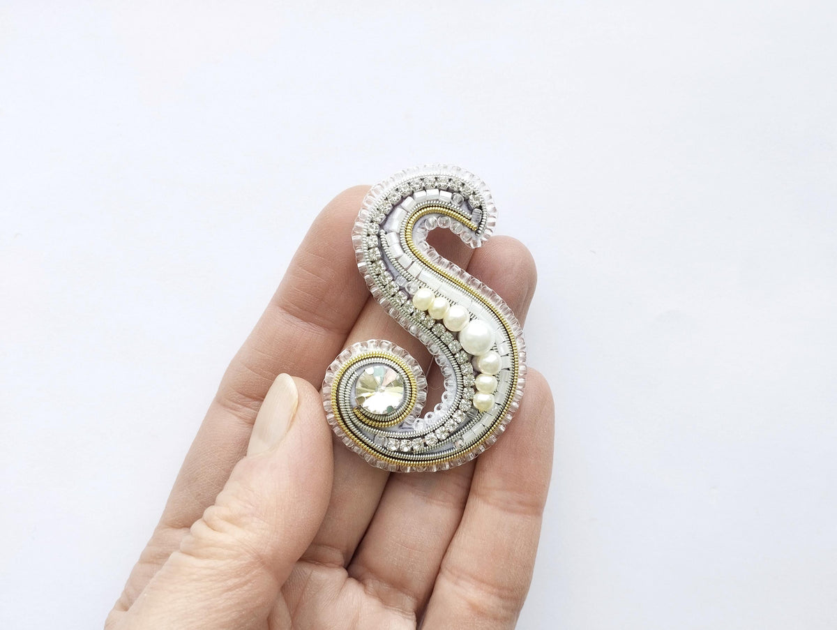A hand holding a decorative letter 'S' made of various materials, including pearls and gemstones.