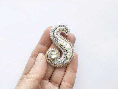 A hand holding a decorative letter 'S' made of various materials, including pearls and gemstones.