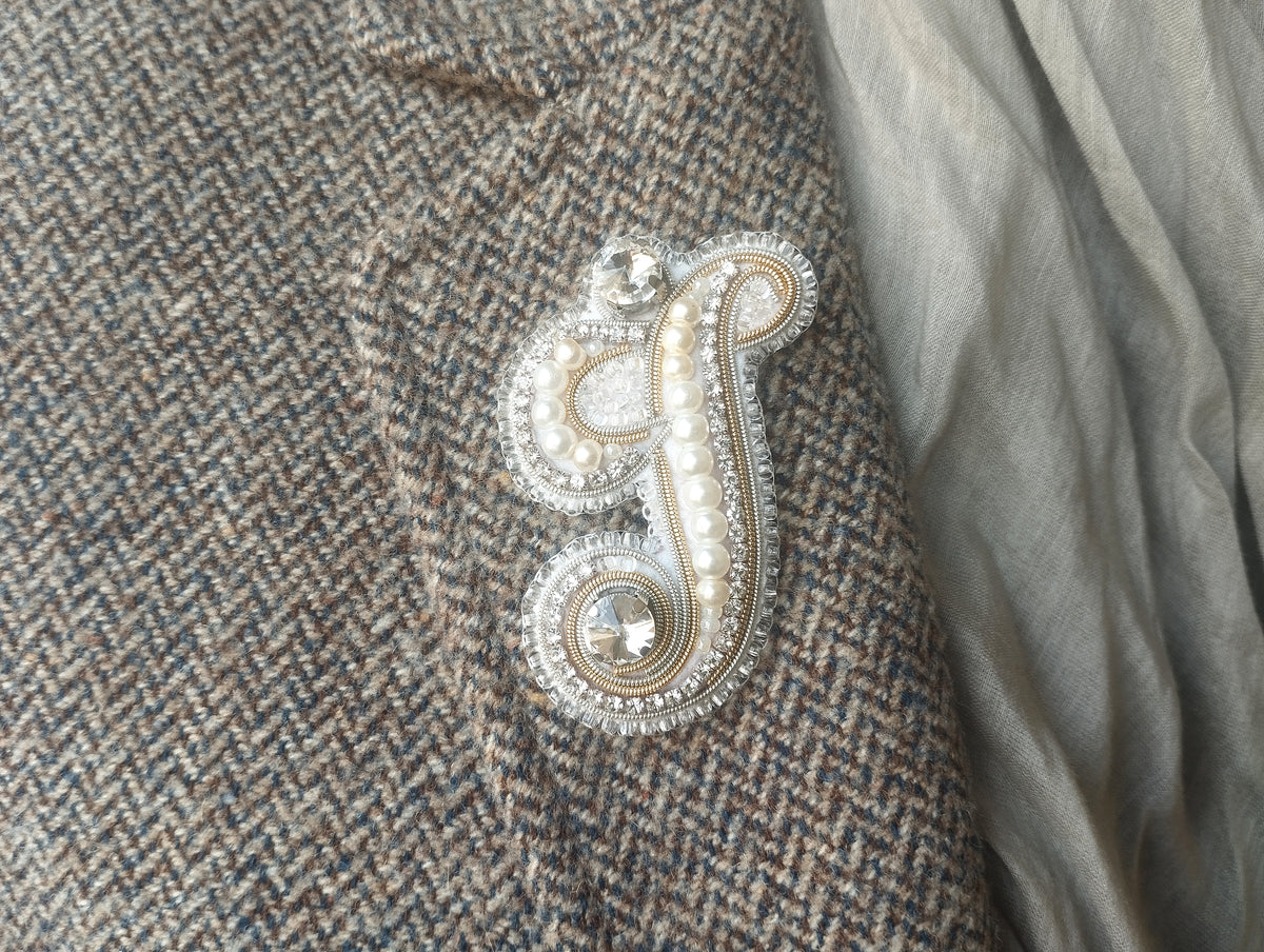 a close-up view of a fabric with a decorative letter "S" made of beads and pearls, placed on a grey fabric.