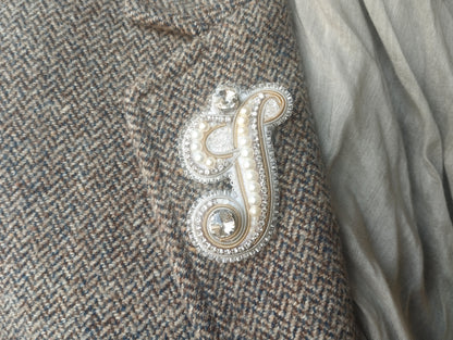a close-up view of a fabric with a decorative letter "S" made of beads and pearls, placed on a grey fabric.