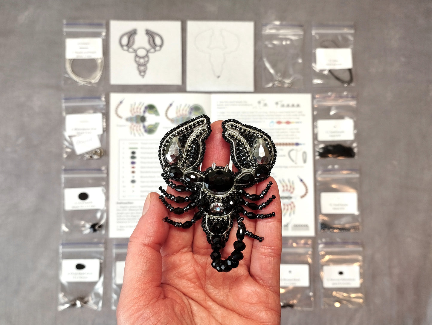 A hand holding a black and silver decorative piece, possibly a brooch or necklace, in front of a display of various jewelry items and materials.