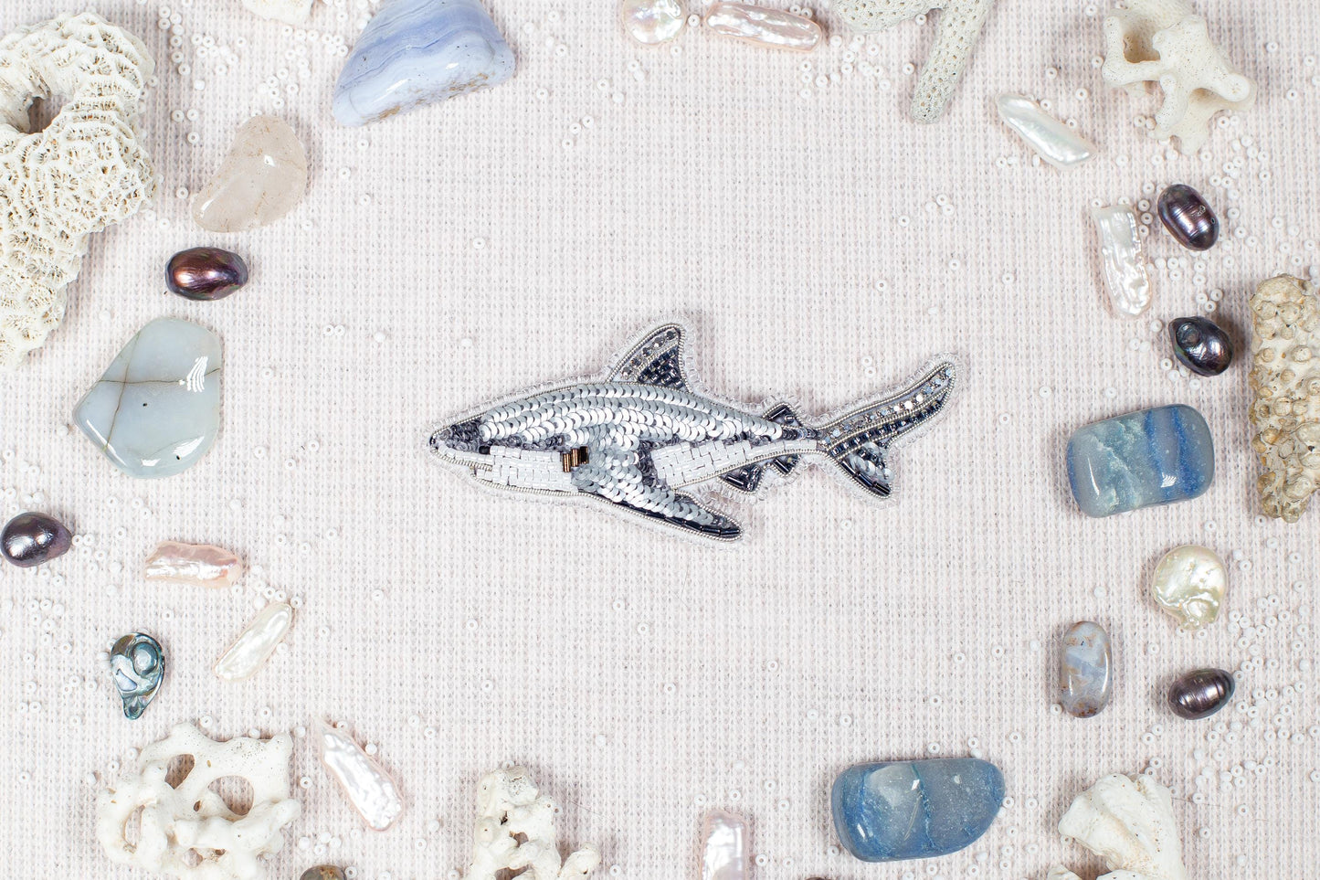 a silver shark embroidered on a light pink background, surrounded by various seashells, pearls, and crystals.