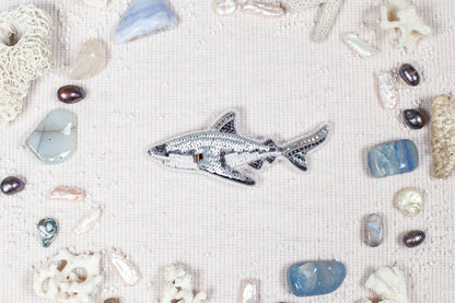 a silver shark embroidered on a light pink background, surrounded by various seashells, pearls, and crystals.