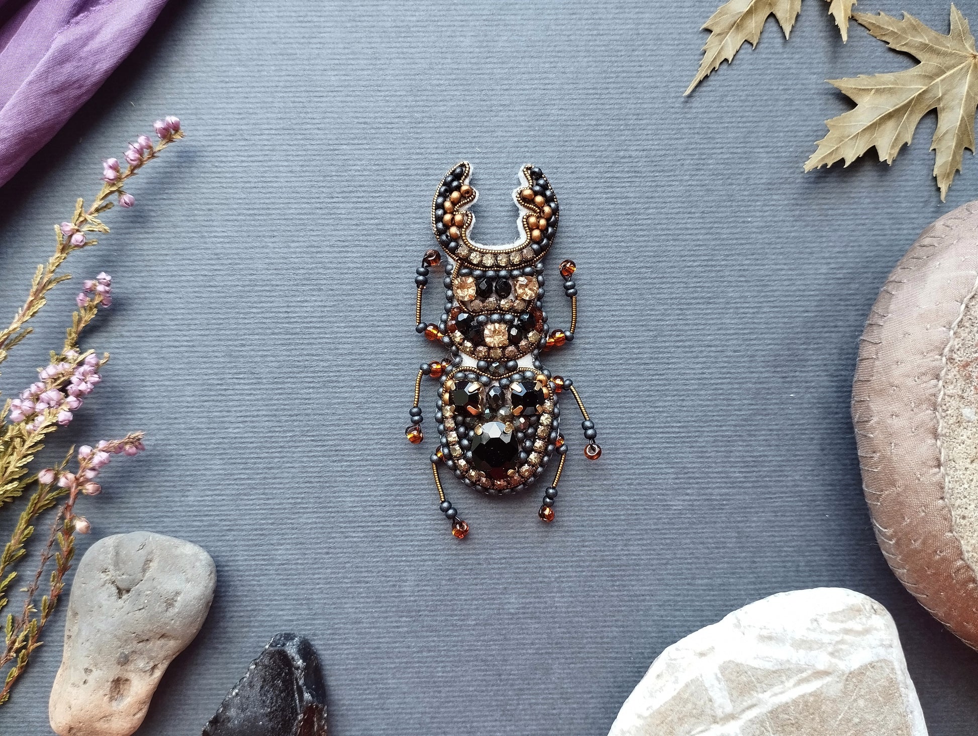 a detailed embroidered or beaded insect, possibly a beetle, set against a grey background with various natural elements such as leaves, rocks, and flowers.