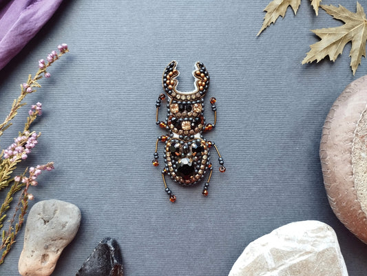 a detailed embroidered or beaded insect, possibly a beetle, set against a grey background with various natural elements such as leaves, rocks, and flowers.