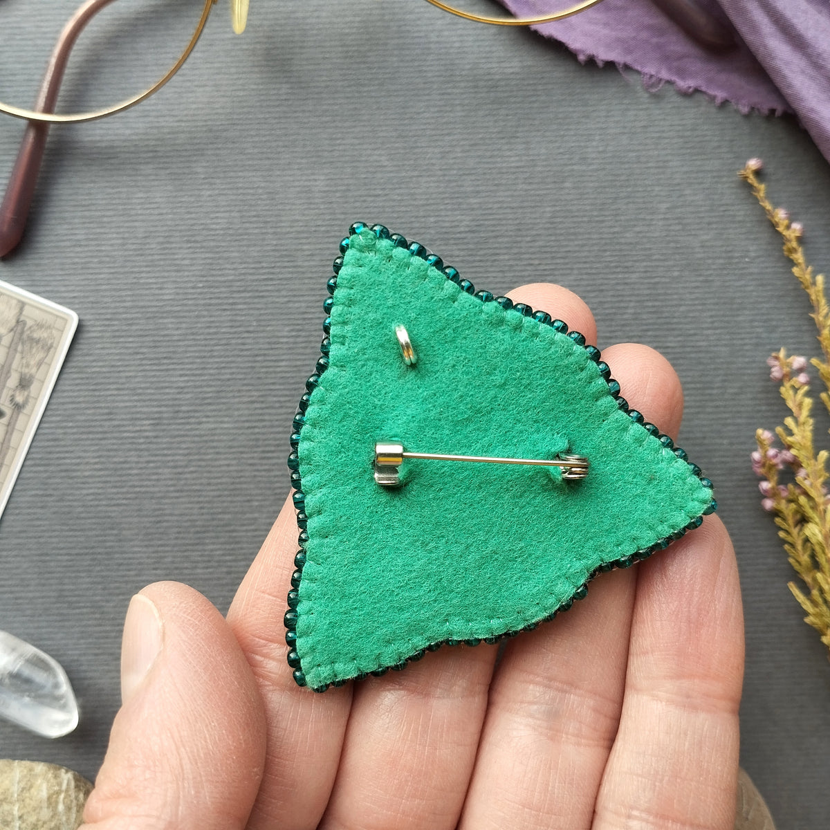 A hand holding a green felt pin with a silver pin attached to it, placed on a gray surface.