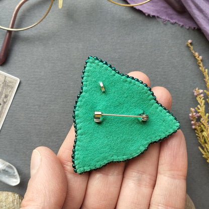 A hand holding a green felt pin with a silver pin attached to it, placed on a gray surface.