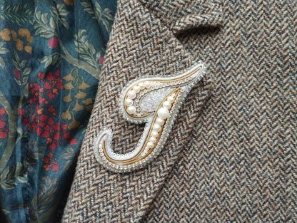 a close-up view of a decorative monogram brooch on a tweed jacket, with a floral patterned fabric visible in the background.