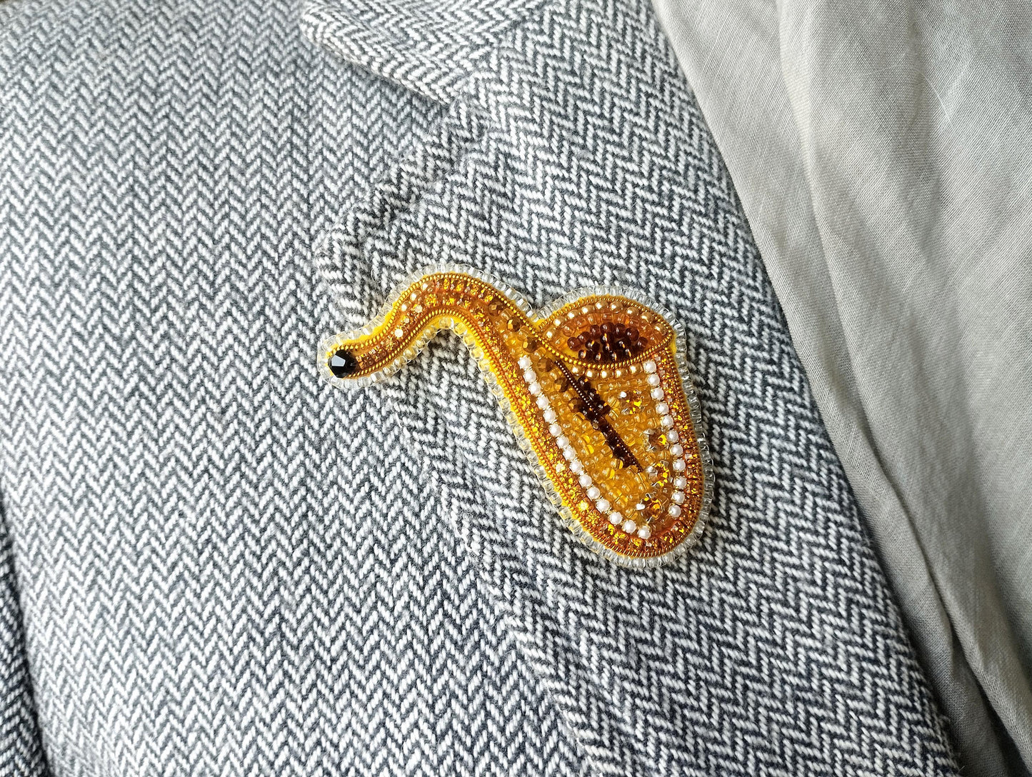 A close-up image of a gold and yellow beaded brooch shaped like a saxophone, attached to a gray and white checkered jacket.