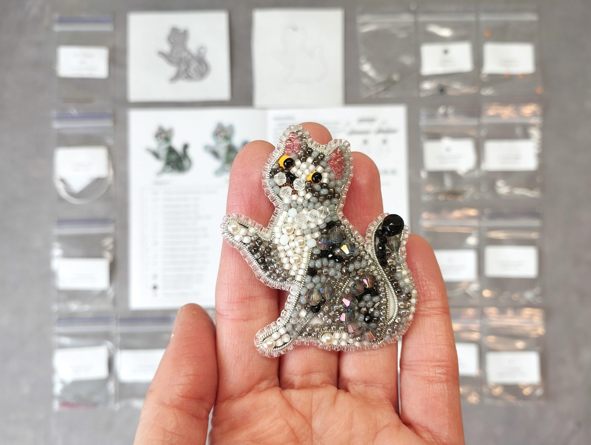 A hand holding a small, intricately beaded cat-shaped brooch against a background of various craft supplies and materials.