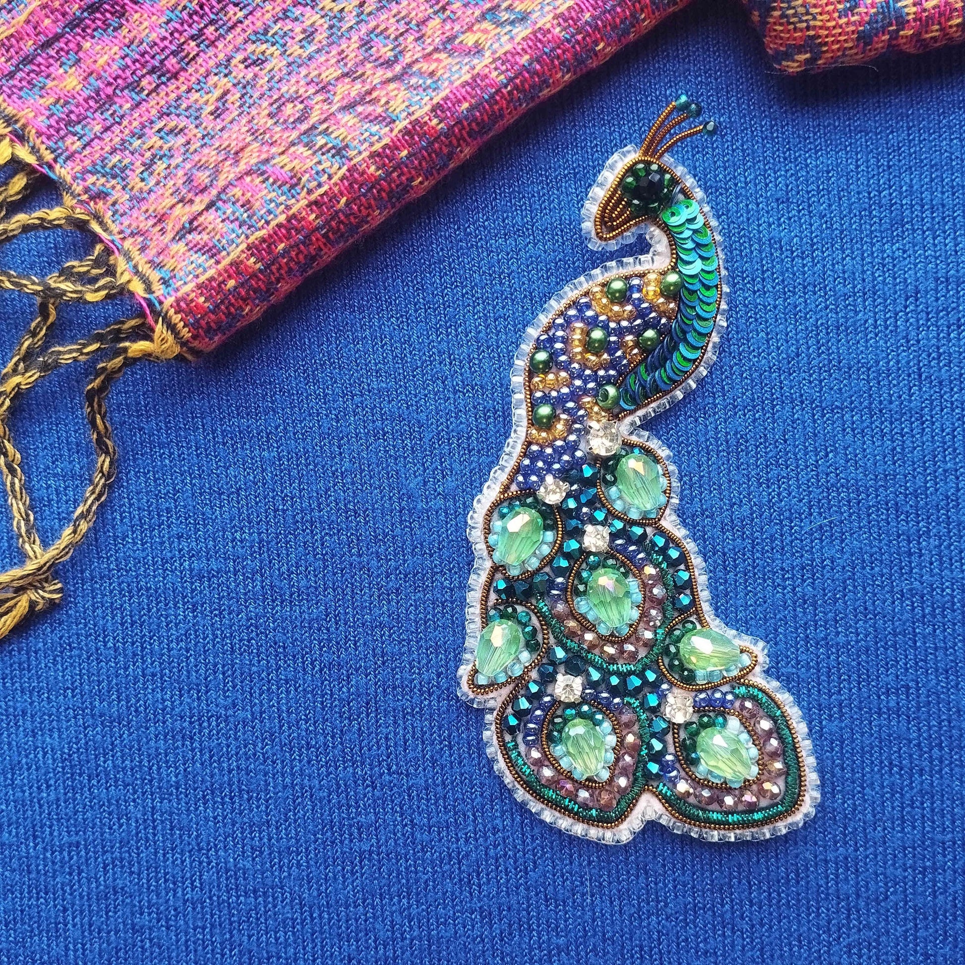 A colorful, intricately designed peacock feather brooch is displayed on a blue fabric surface, with a multicolored woven fabric visible in the background.