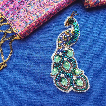 A colorful, intricately designed peacock feather brooch is displayed on a blue fabric surface, with a multicolored woven fabric visible in the background.