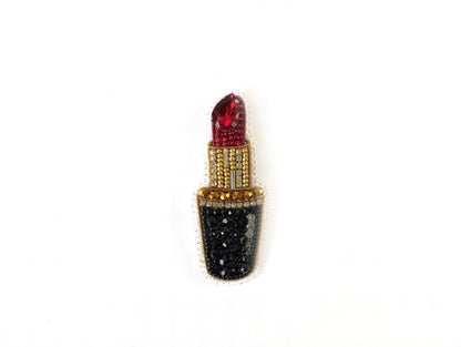 A decorative piece featuring a red lipstick with a gold and black design, including rhinestones and a gemstone.