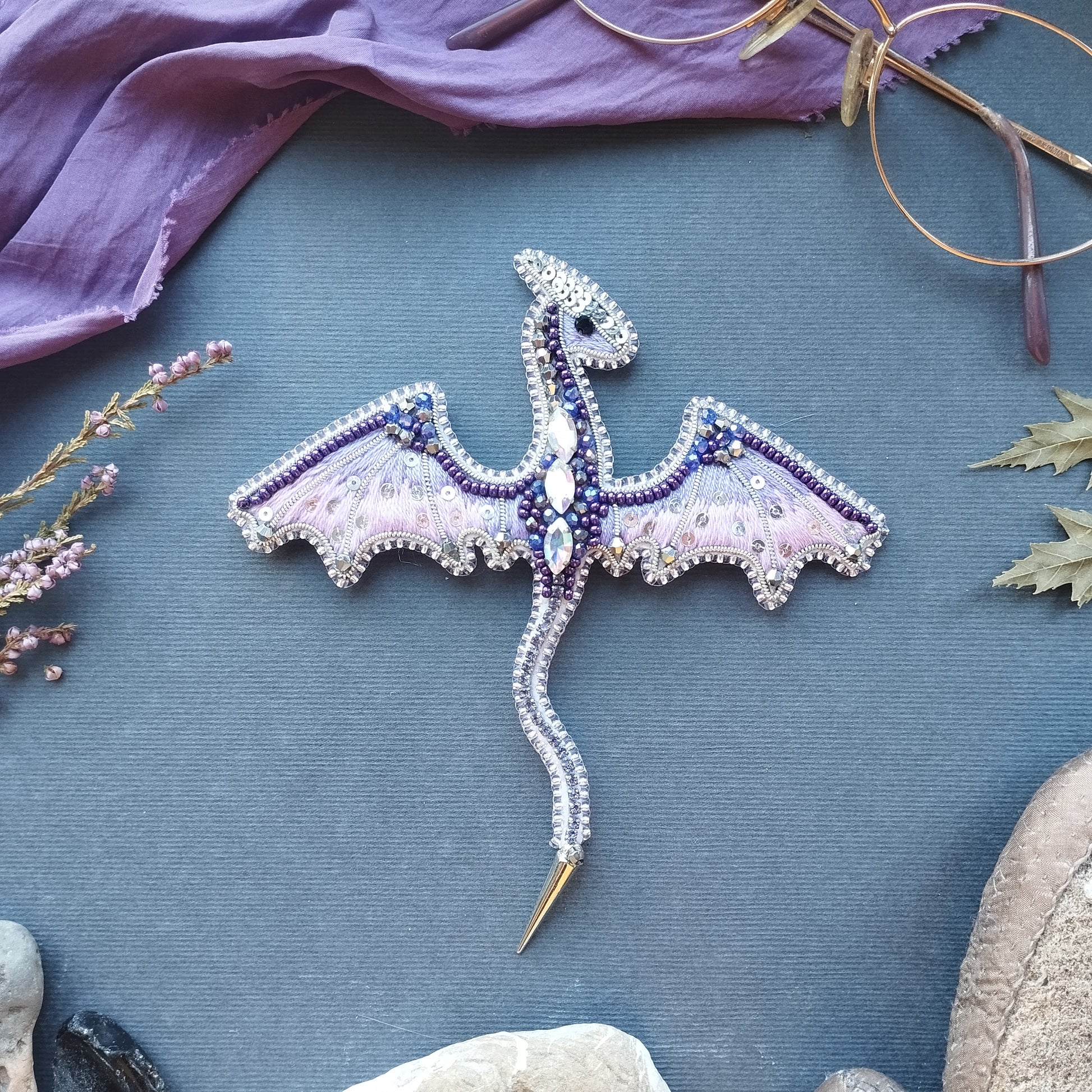 a decorative dragon brooch with intricate beading and gemstone details, placed on a blue background alongside other items such as a pair of glasses, a purple cloth, and some rocks.