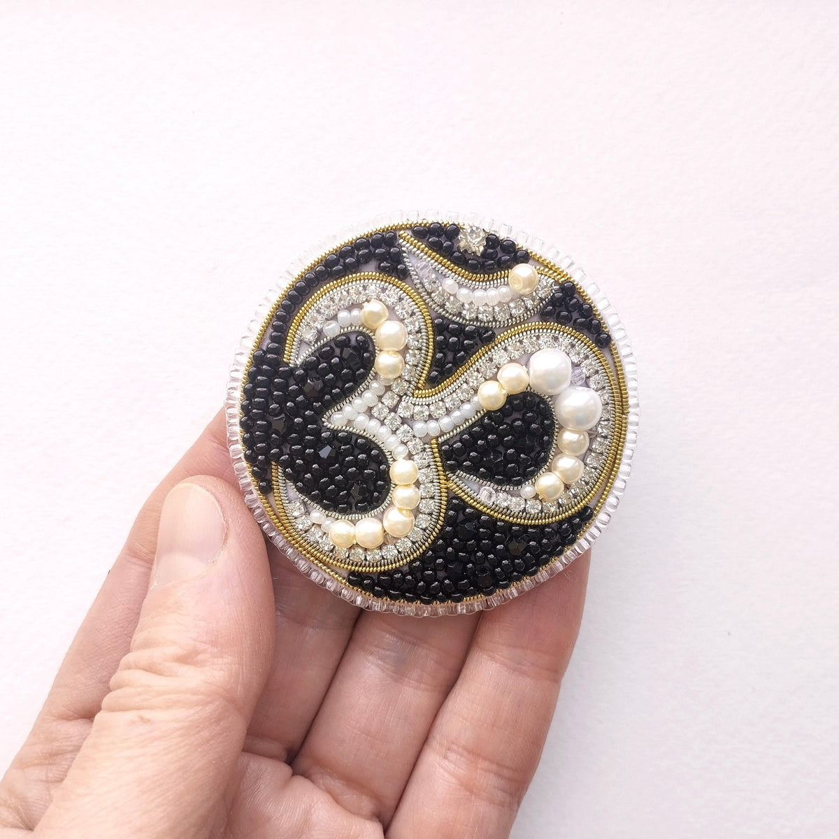 A hand holding a circular, ornate piece of jewelry with a black and white design, including pearls and beads.