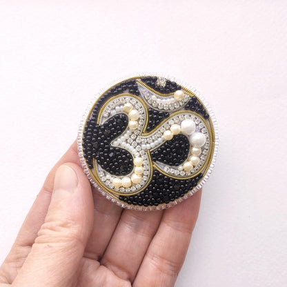A hand holding a circular, ornate piece of jewelry with a black and white design, including pearls and beads.