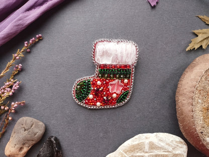 A decorative Christmas stocking adorned with colorful beads and a white trim is displayed on a gray background, surrounded by various natural elements such as leaves, rocks, and a purple fabric.