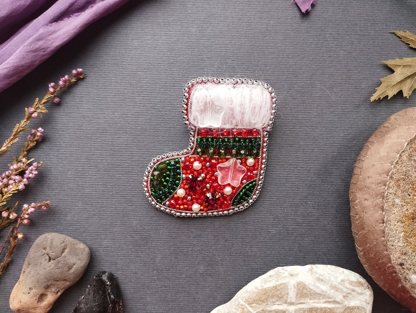 A decorative Christmas stocking adorned with colorful beads and a white trim is displayed on a gray background, surrounded by various natural elements such as leaves, rocks, and a purple fabric.