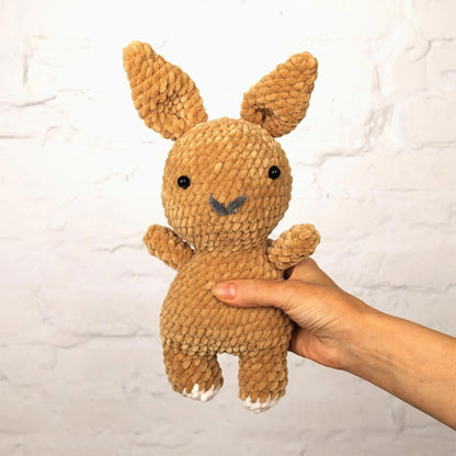 A hand is holding a brown, crocheted stuffed rabbit toy against a white brick wall background.