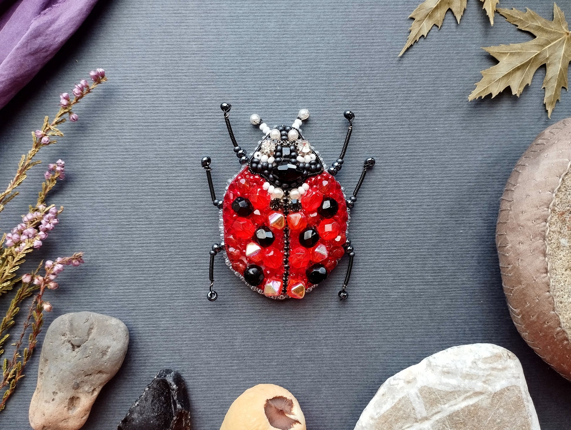 A red ladybug-shaped beaded object is displayed on a gray background, surrounded by various natural elements such as leaves, rocks, and flowers.